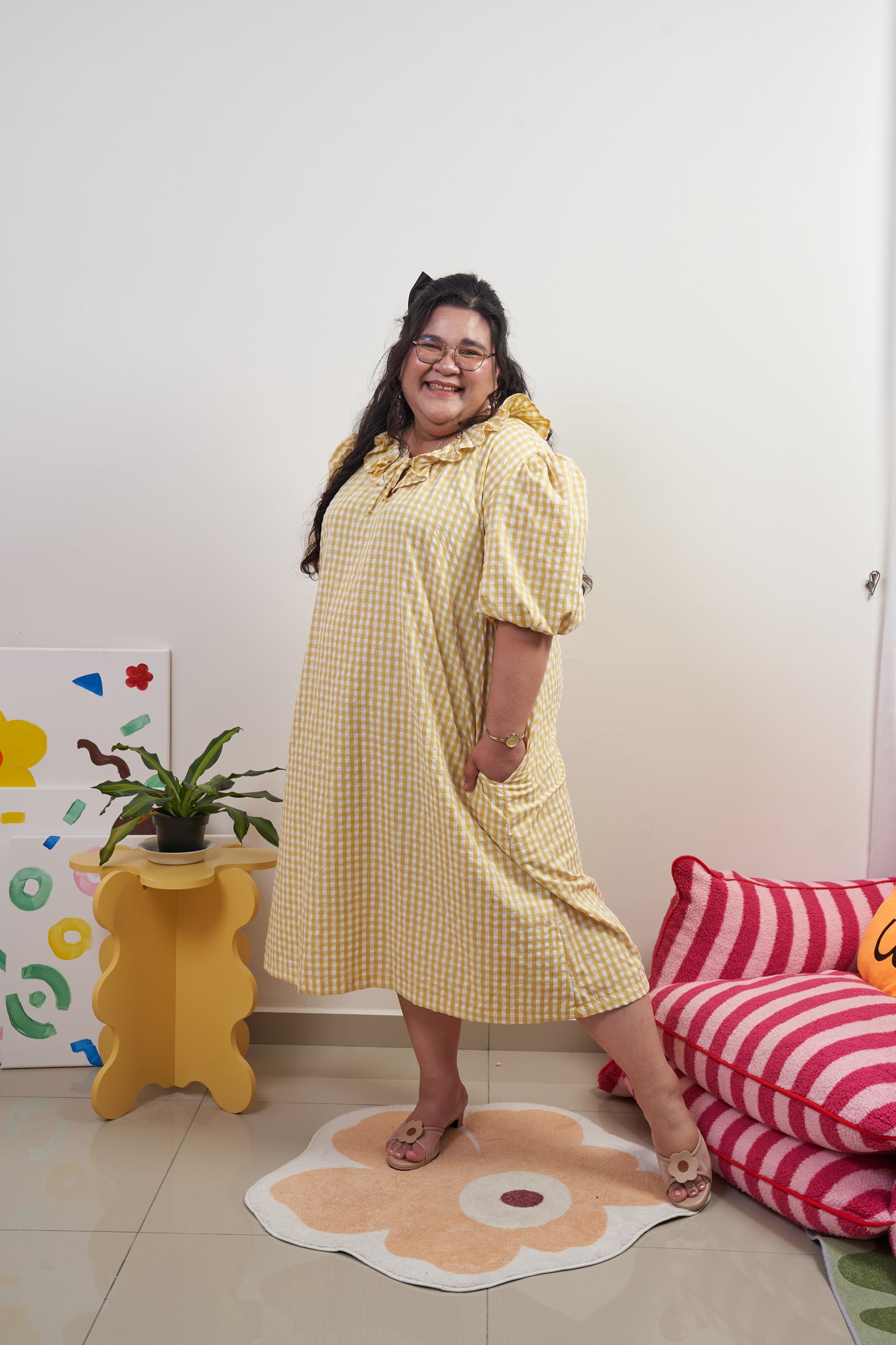 Full-body side-front view of Bea Dress in Sunny Gingham, model with one hand in pocket, featuring yellow gingham print, puffed sleeves, adjustable ruffled neckline, and relaxed trapeze silhouette
