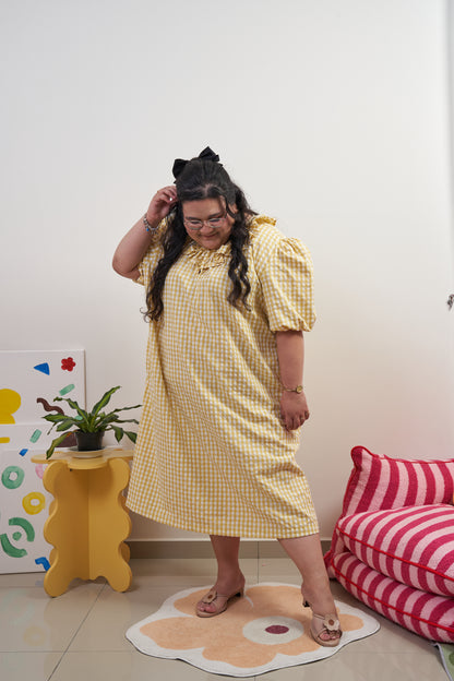 Side-profile view of Bea Dress in Sunny Gingham, model in relaxed pose with one leg slightly forward, showcasing yellow gingham print, puffed sleeves, adjustable ruffled neckline, and trapeze silhouette