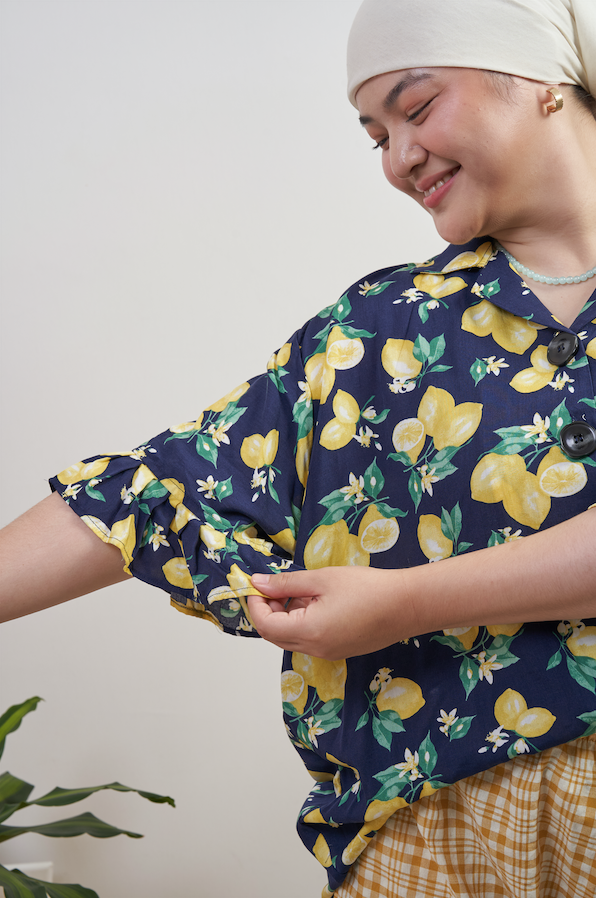 Semi-body close-up of Buttoned Belle Blouse in Lemon Dream, model admiring ruffled sleeve detail with joyful expression, styled with Flutter Flair Pants in Lemonade.