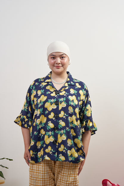 Model smiling and relaxed in Buttoned Belle Blouse in Lemon Dream, showing vintage buttons and ruffled sleeves against a dark blue base with lemon print.