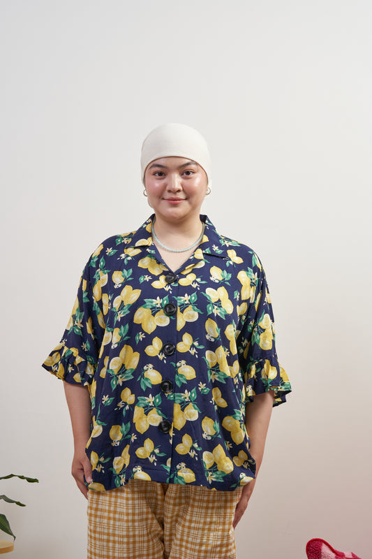 Model smiling and relaxed in Buttoned Belle Blouse in Lemon Dream, showing vintage buttons and ruffled sleeves against a dark blue base with lemon print.