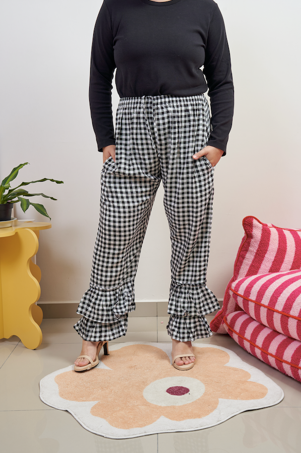 Front view of Flutter Flair Pants in Noir Blanc, model with hands in pockets, showcasing black & white gingham pattern, double ruffle hem, drawstring waist, and loose fit