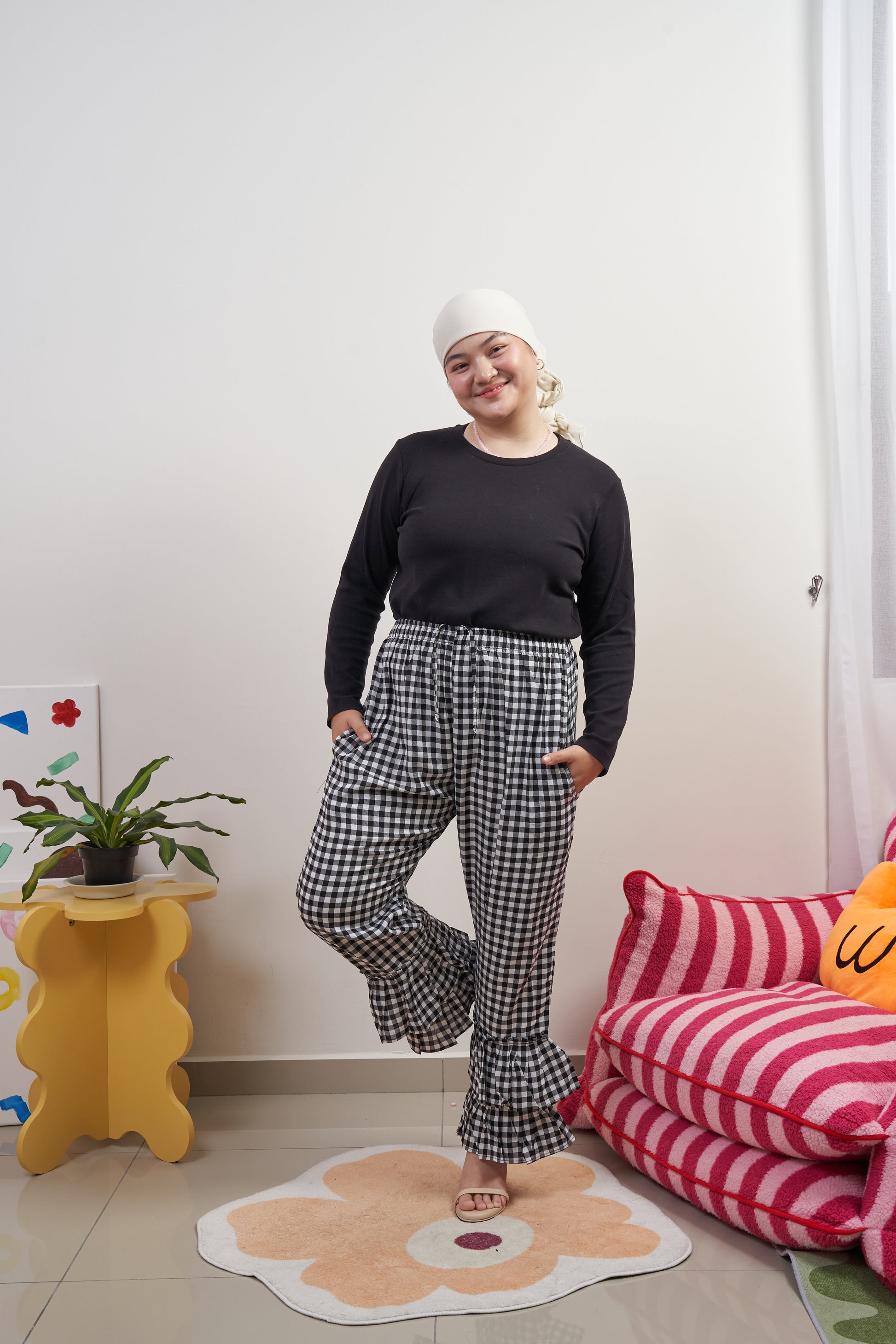 Full-body view of Flutter Flair Pants in Noir Blanc, model in relaxed stance with one leg lifted back, showcasing black & white gingham, double ruffle hem, drawstring waist, and side pockets.