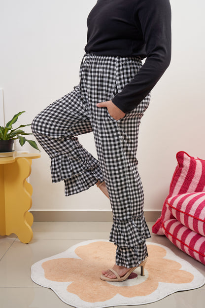 Side profile of Flutter Flair Pants in Noir Blanc, model lifting right leg forward with one hand in pocket, showcasing black & white gingham, double ruffle hem, and loose fit.