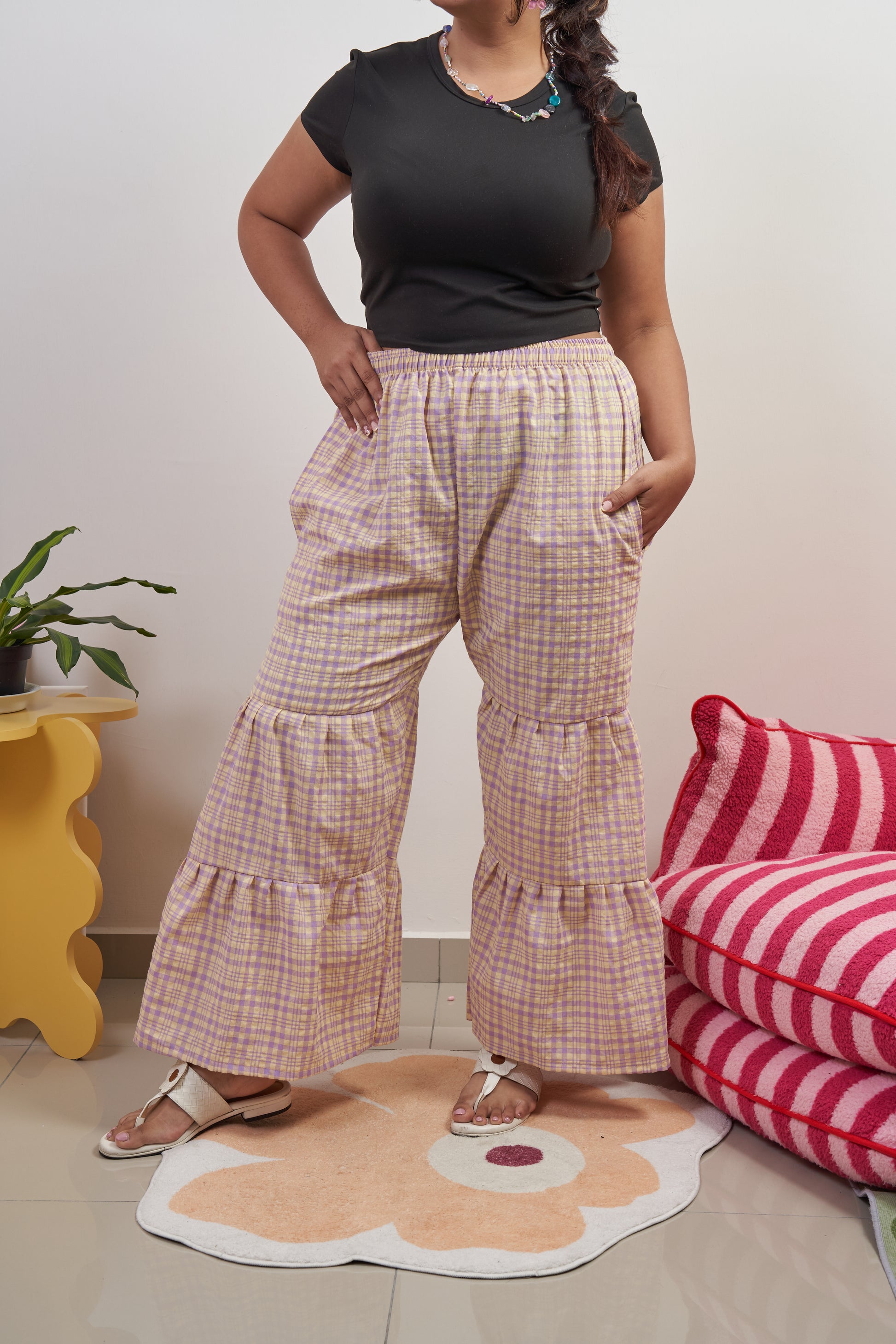 ront profile of Flutter Tier Pants in Orchid Zest, model with one leg extended to showcase purple and yellow plaid pattern, double-tier ruffled flared hem, and loose fit.