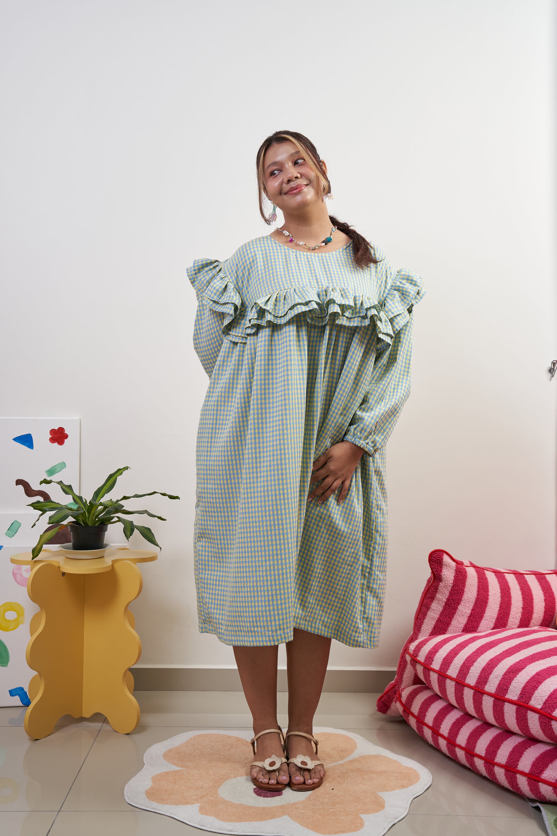 Semi-frontal view of Rhea Dress in Felicia Daisy, model leaning forward for an elegant and demure look, featuring yellow and blue gingham print with a flowing trapeze silhouette