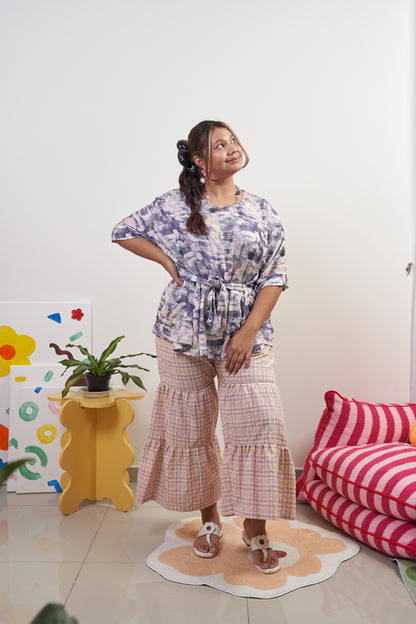 Full-body angled view of Liliana Top in Wisteria with ribbon tied at the front, paired with Flutter Tier Pants in Orchid Zest, showing relaxed, breezy fit and peplum style.