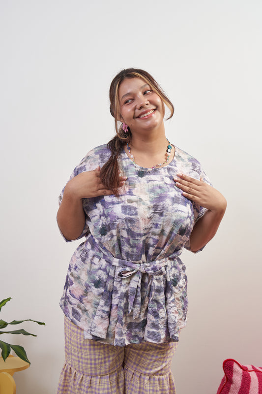 Front view of Liliana Top in Wisteria with ribbon tied in front, showcasing soft purple peplum style, round neckline, wide cuff sleeves, and ruffled hem.