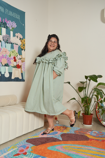 Model lifting one leg in a playful jump pose wearing the Rhea Dress in Felicia Daisy, showcasing yellow and blue gingham print with ruffles and flowing tent dress silhouette