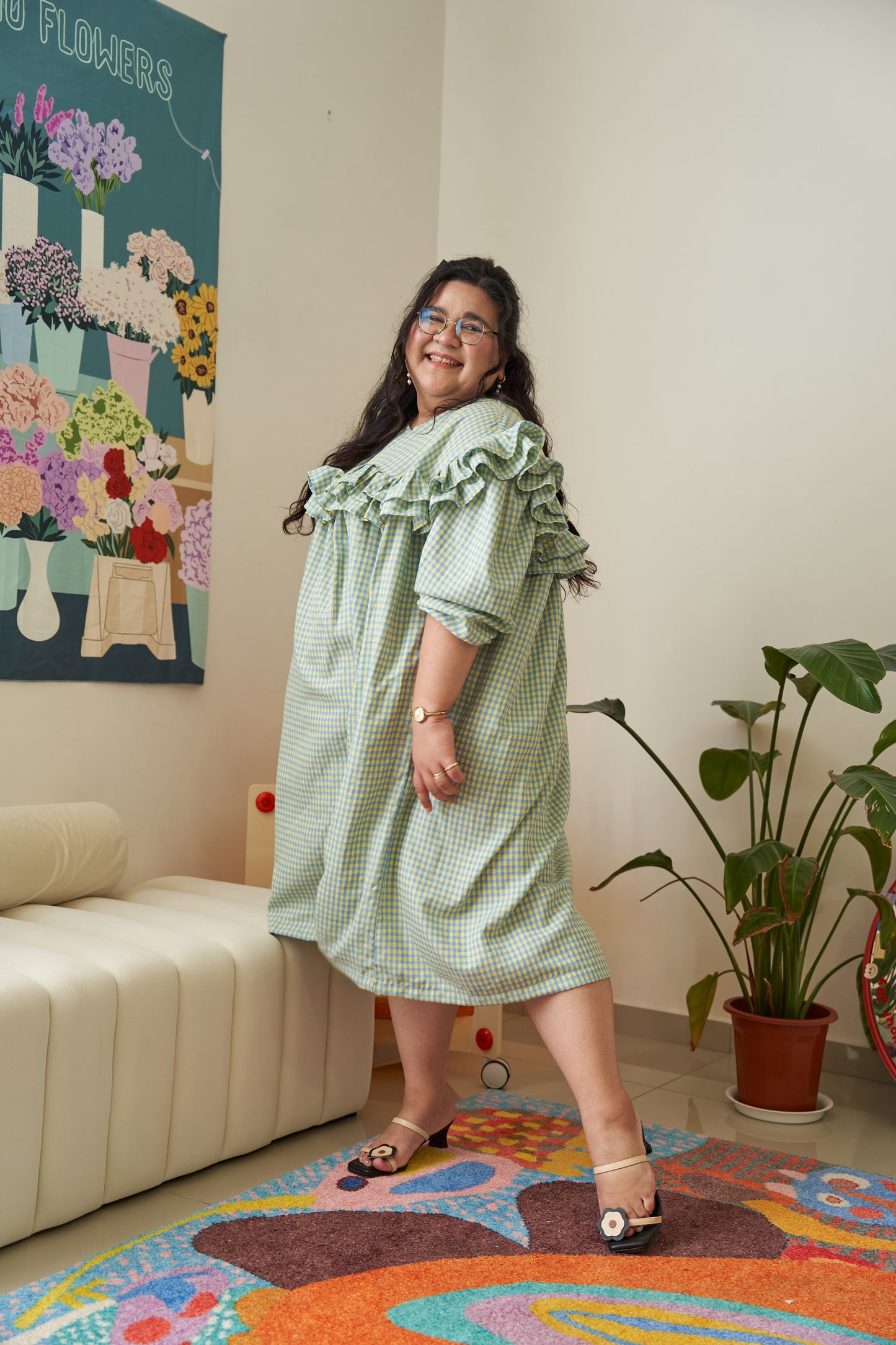 Semi-side frontal view of Rhea Dress in Felicia Daisy, model in a different pose, featuring yellow and blue gingham print with ruffles and a flowing tent dress silhouette