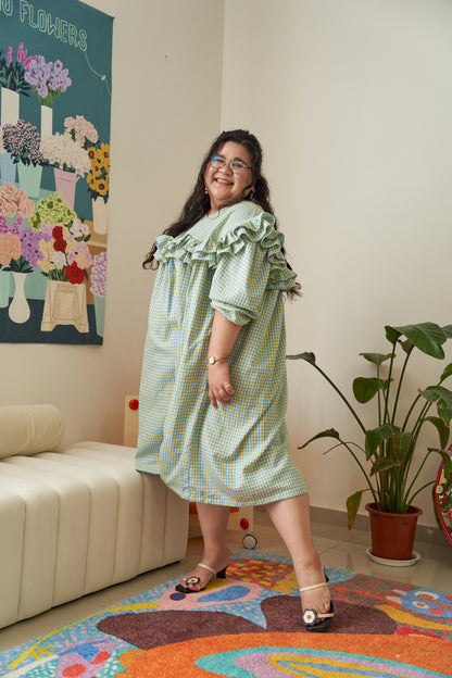 Semi-side frontal view of Rhea Dress in Felicia Daisy, model in a different pose, featuring yellow and blue gingham print with ruffles and a flowing tent dress silhouette