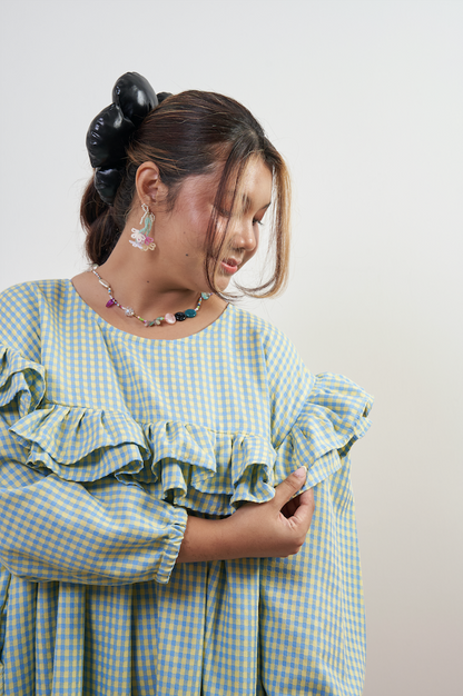 Half-body side view of Rhea Dress in Felicia Daisy, model holding the ruffle to showcase elegance and demure style, with yellow and blue gingham print