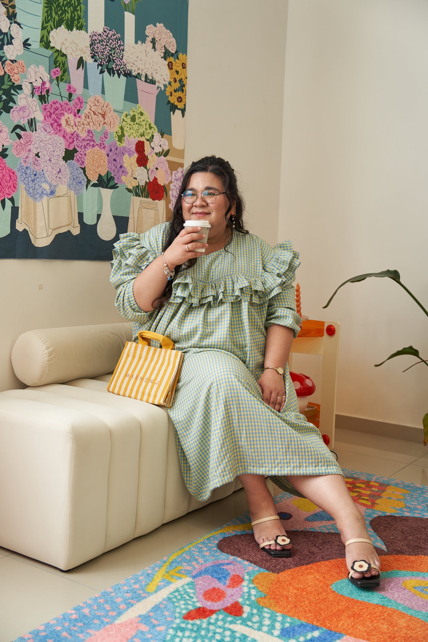 Model sitting down and drinking coffee while wearing the Rhea Dress in Felicia Daisy, featuring yellow and blue gingham print with ruffles and a relaxed tent dress silhouette