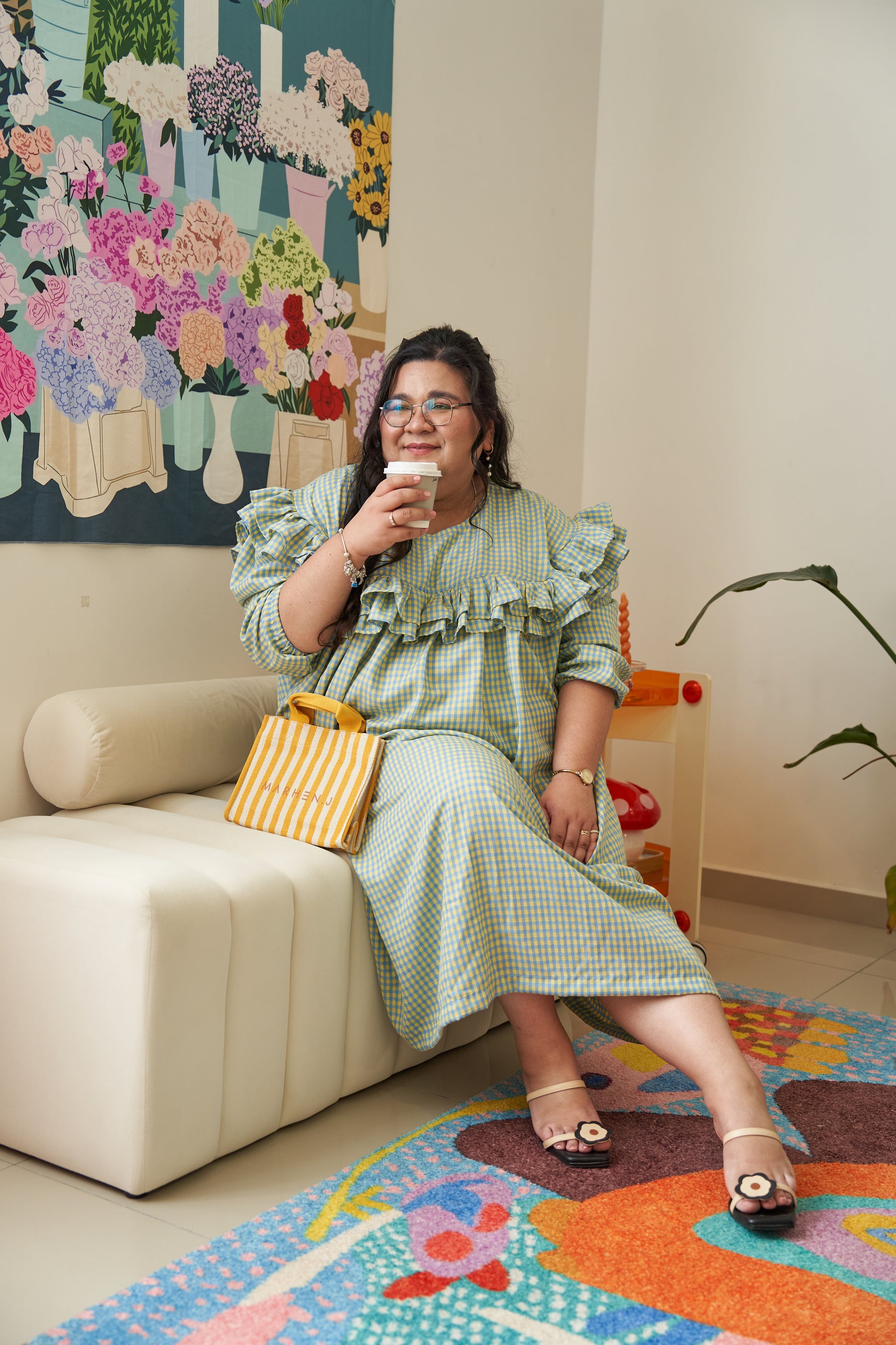 Model sitting down and drinking coffee while wearing the Rhea Dress in Felicia Daisy, featuring yellow and blue gingham print with ruffles and a relaxed tent dress silhouette