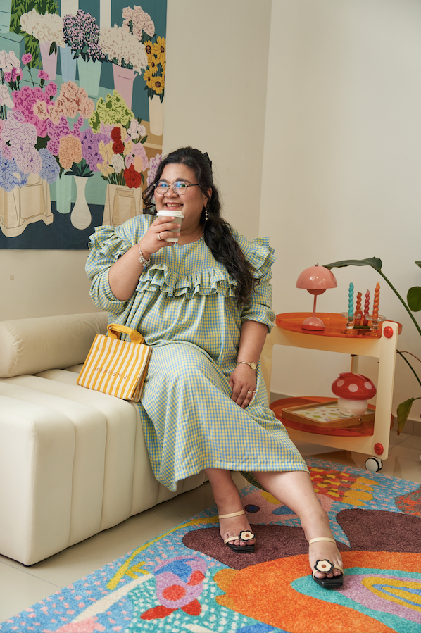 Model sitting down facing the camera and drinking coffee, wearing the Rhea Dress in Felicia Daisy, with yellow and blue gingham print and ruffles in a relaxed tent dress silhouette