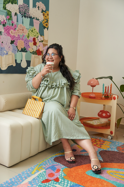 Model sitting down facing the camera and drinking coffee, wearing the Rhea Dress in Felicia Daisy, with yellow and blue gingham print and ruffles in a relaxed tent dress silhouette