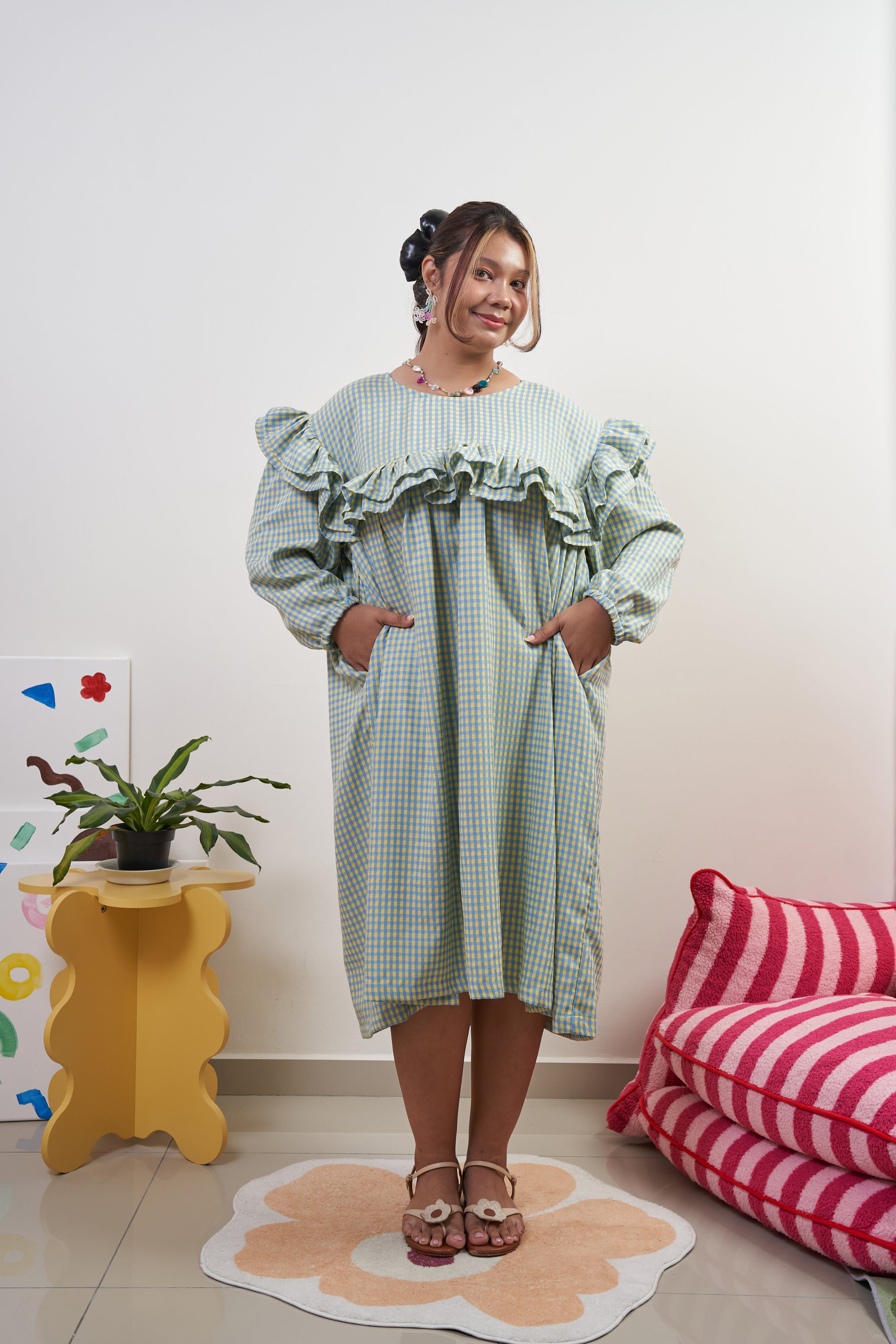 Front view of Rhea Dress in Felicia Daisy with model’s hands in pockets, yellow and blue gingham print, featuring trapeze silhouette and ruffles